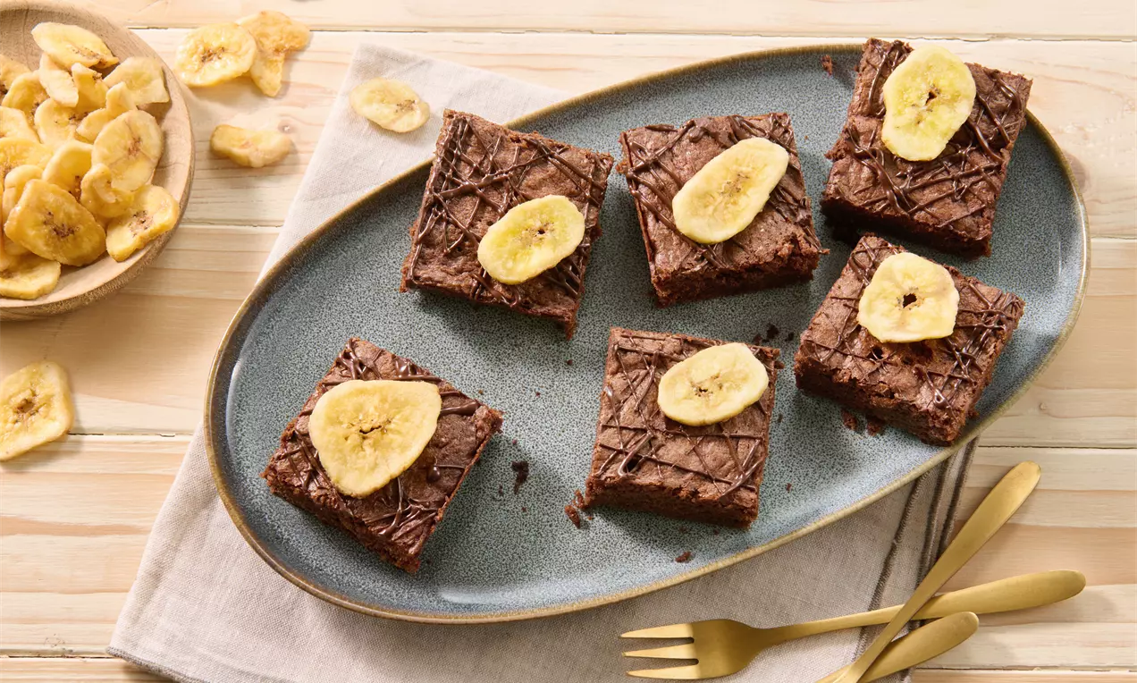 Saftige, quadratische Schoko-Brownies mit glatter Oberfläche, einzelnen getrockneten Bananenscheiben garniert auf blau-grauer Platte angerichtet