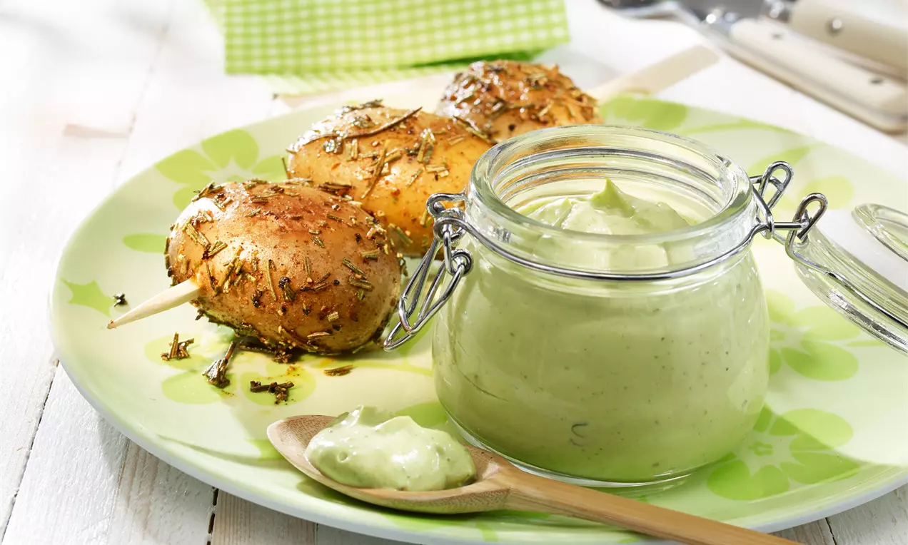 Goldbraune Kartoffelspieße mit Kräutern auf grünem Teller neben cremigem Avocado-Basilikum-Dip in einem Gläschen und Holzlöffel