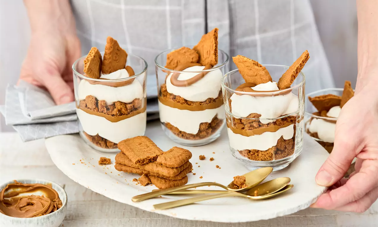 Veganes Lotus-Dessert in drei Gläsern, geschichtet mit Keksstücken, Creme und Lotus-Aufstrich. Garniert mit ganzen Keksen. Daneben goldene Löffel, extra Kekse und eine Schale mit Aufstrich.