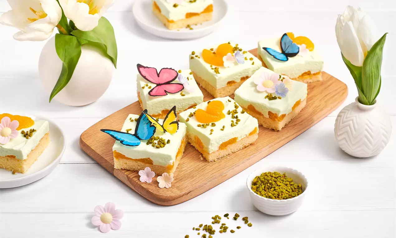Frühlingskuchen mit hellgrüner Creme auf gelbem Biskuit, dekoriert mit essbaren Schmetterlingen, Blüten und Pistaziensplittern auf Holzbrett