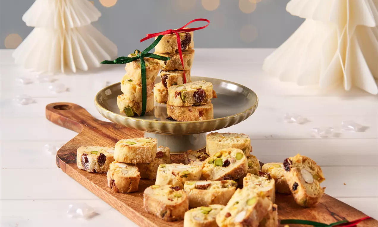 Goldbraune, knusprige Cantuccini mit Pistazien und Cranberries, auf Holzbrett und Teller arrangiert, mit roten und grünen Bändern verziert