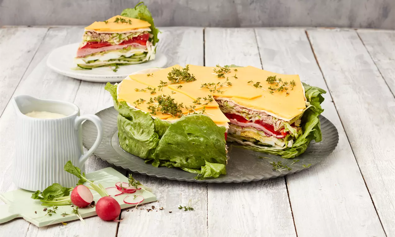 Salattorte mit Schichten aus grünem Salat, Schinken, Käse und roten Tomaten, garniert mit Kräutern auf einer Platte