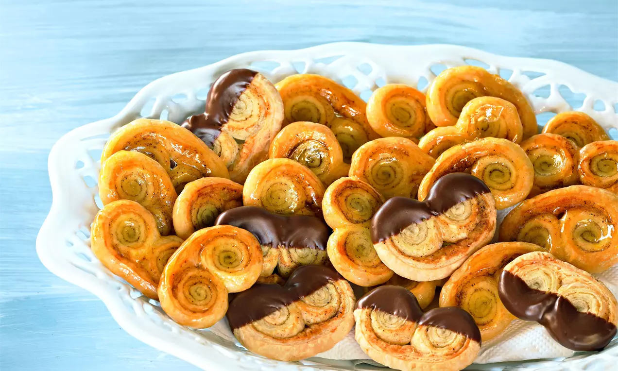 Goldbraune, spiralige Schweineöhrchen mit knuspriger Textur und teils dunkler Schokoladenhälfte auf weißem Spitzenteller angerichtet