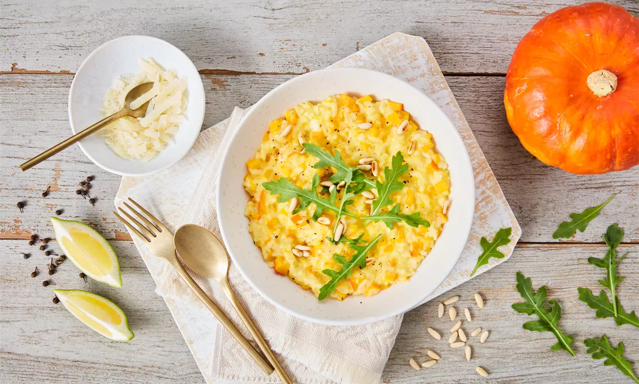 Cremiges, goldgelbes Kürbisrisotto in einer weißen Schale, garniert mit Rucola und Kernen, angerichtet auf einem Holzuntergrund mit Dekoration
