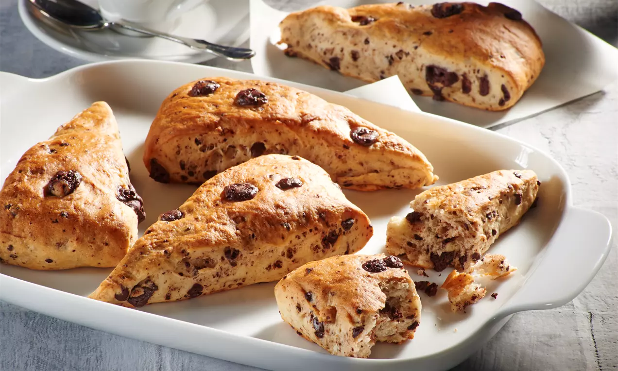 Goldbraune, dreieckige vegane Schokobrötchen mit dunklen Schokostückchen auf einer weißen Keramikplatte, teils gebrochen und locker