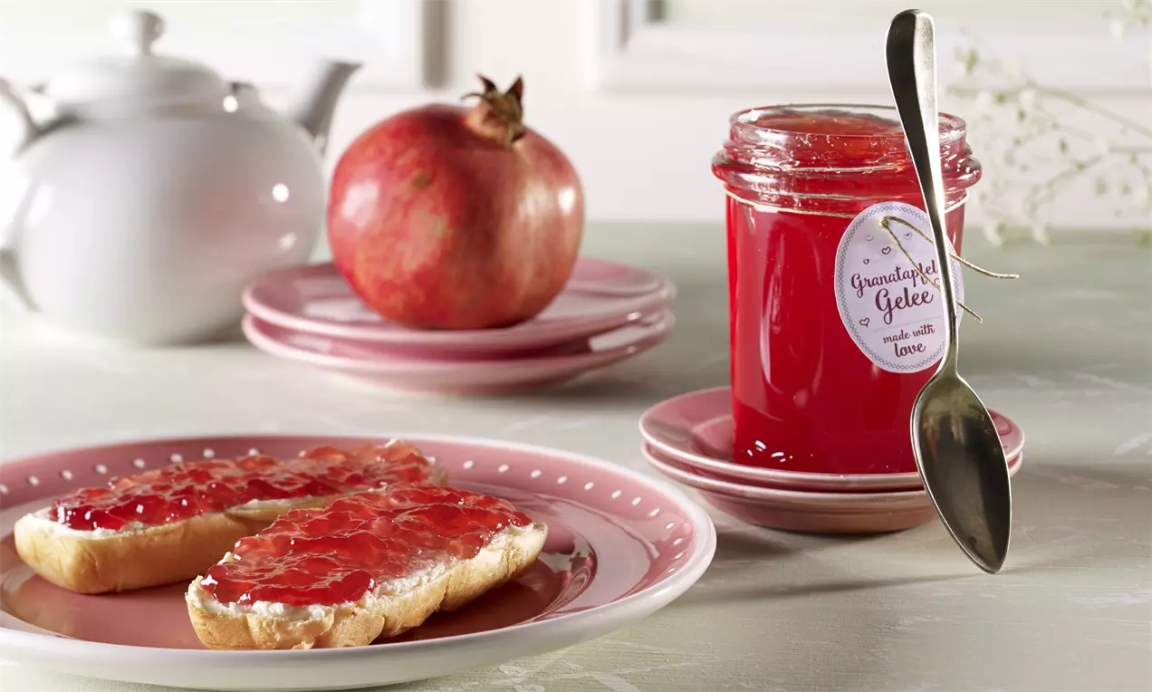 Leuchtend rotes Apfel-Granatapfel-Gelee in einem Glas neben aufgeschnittenem Brötchen mit Gelee und fruchtiger Granatapfeldeko auf rosa Tellern