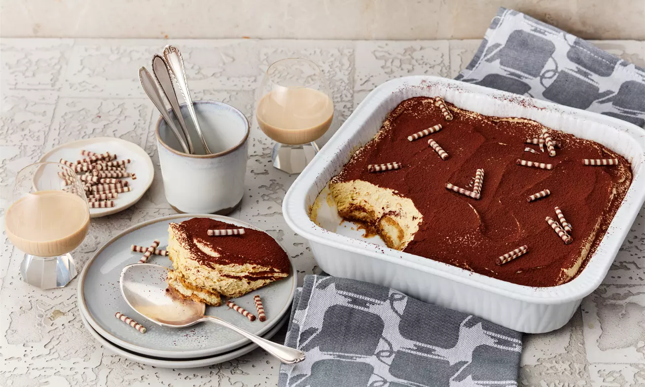 Baileys-Tiramisu mit hellgelber Creme, Kakaopulver und eingerollter Deko in weißer Form, daneben ein Teller mit serviertem Stück
