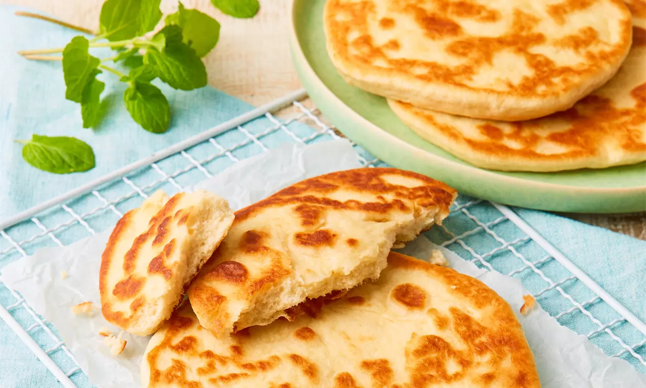 Goldbraunes Pfannenbrot mit weicher, leicht luftiger Textur, gestapelt und teils aufgebrochen auf hellem Pergamentpapier mit Minzzweig