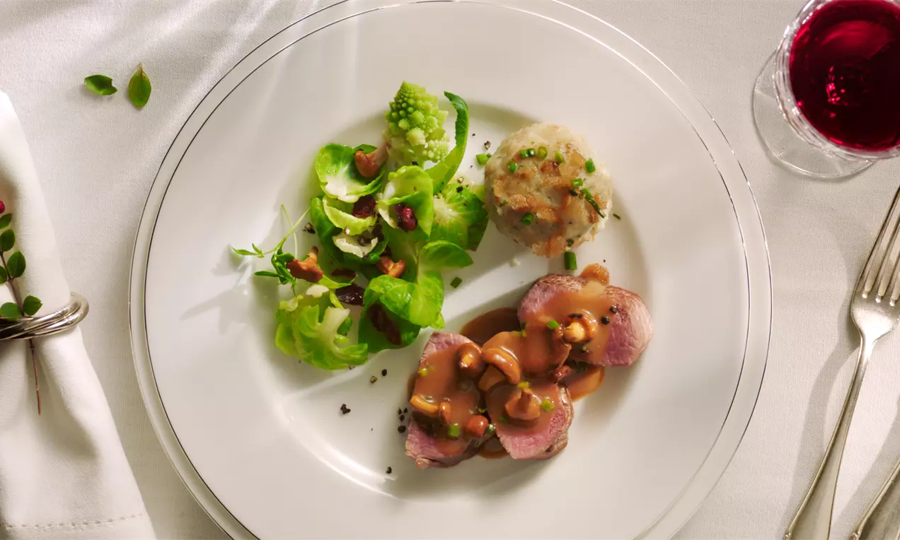 Rosa gebratene Rehrückenmedaillons mit glänzendem braunem Pfifferlingsjus neben grünen Romanesco-Röschen und einem goldbraunen Semmelknödel