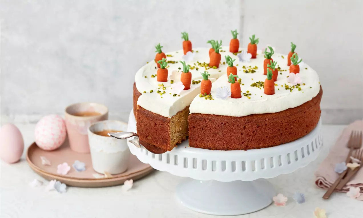 Saftige Möhrentorte mit weißer Cremehaube, dekoriert mit kleinen Marzipanmöhren und Pistazienstückchen auf weißer Tortenplatte
