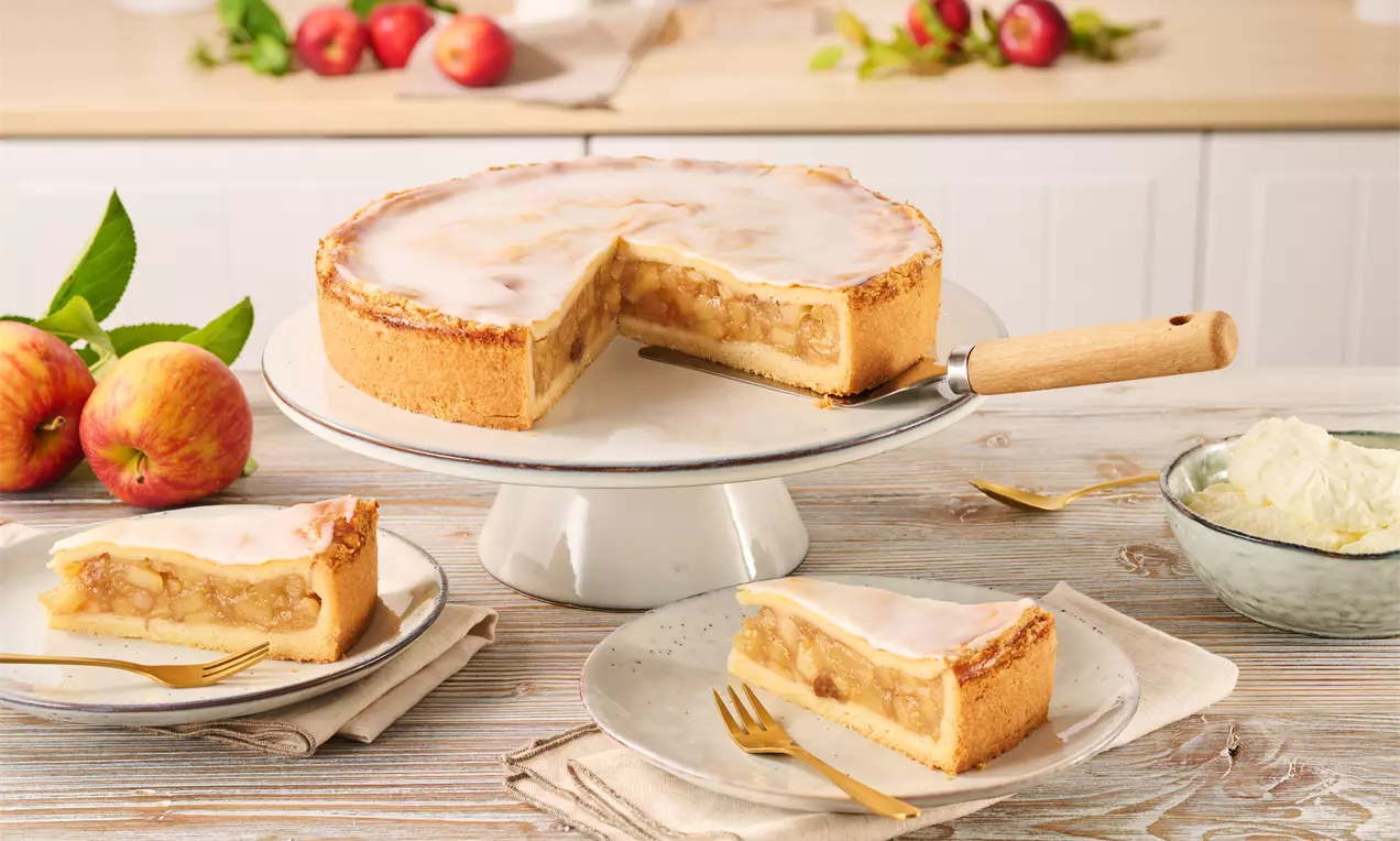 Gedeckter Apfelkuchen mit glänzendem Zuckerguss auf einer Kuchenplatte, zwei Stücke auf Tellern mit Gabeln, daneben frische Äpfel und Sahne