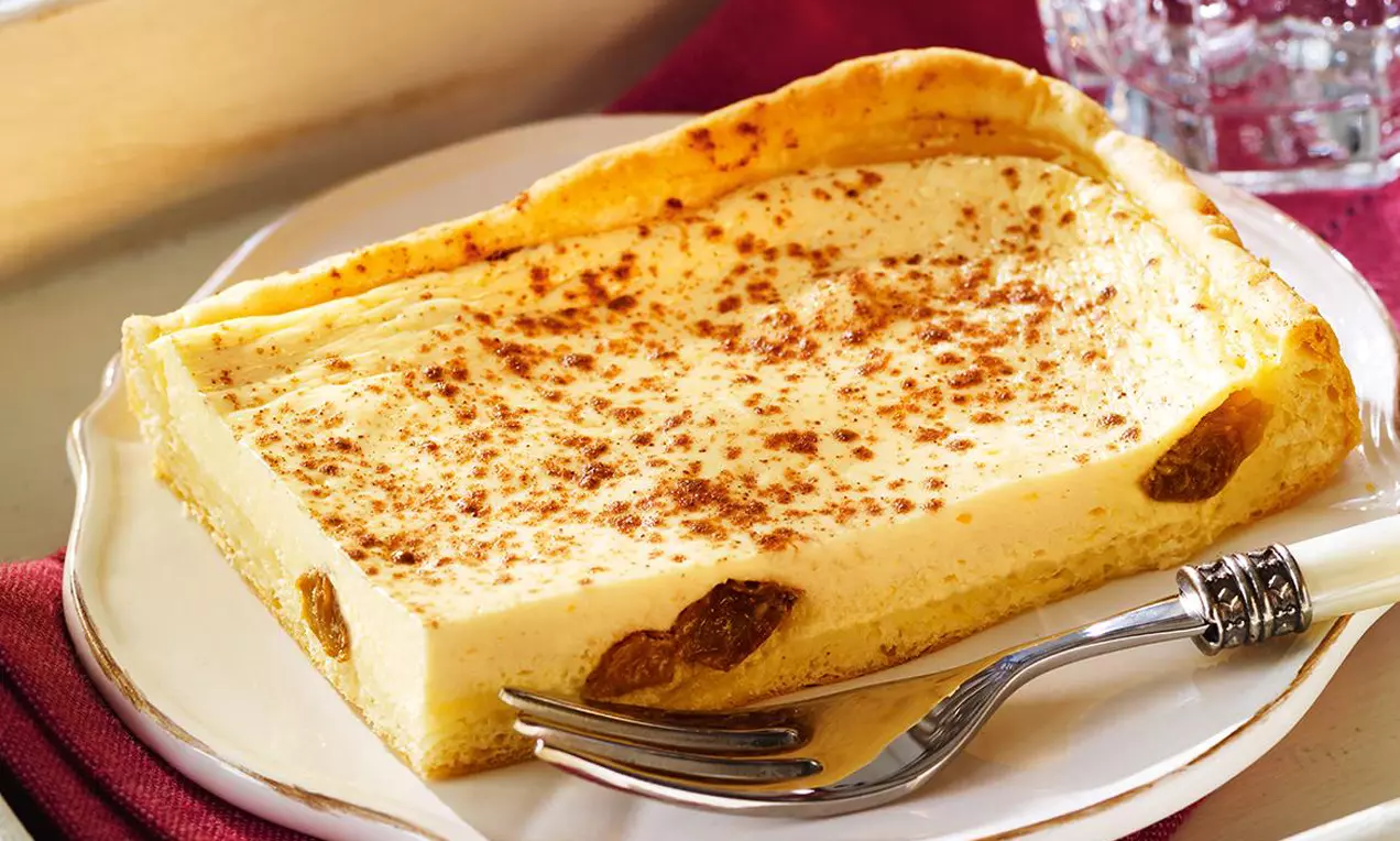 Ein rechteckiges Stück goldgelber Käsekuchen mit feiner Bräunung und Rosinen auf weißem Teller, daneben eine silberne Gabel