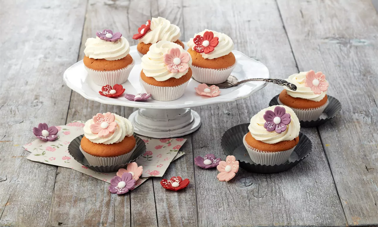 Vanille-Cupcakes mit weißer Sahnehaube, dekoriert mit bunten Blüten aus Fondant, arrangiert auf einer weißen Tortenplatte und Holzuntergrund