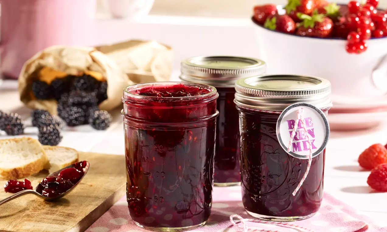 Dunkelrote Konfitüre mit fruchtigen Beerenstücken in klaren Gläsern, umgeben von frischen Beeren, Brot und einem Holzbrett auf rosafarbenem Stoff