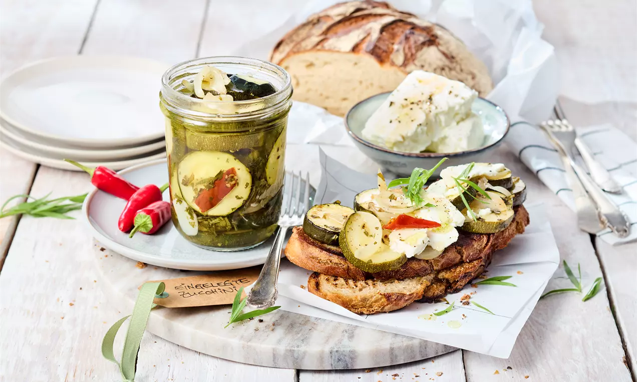 Ein Glas mit grün-gelben Zucchinischeiben in Öl und Gewürzen, daneben Brot, eingelegte Zucchini auf Toast und ein Laib Brot