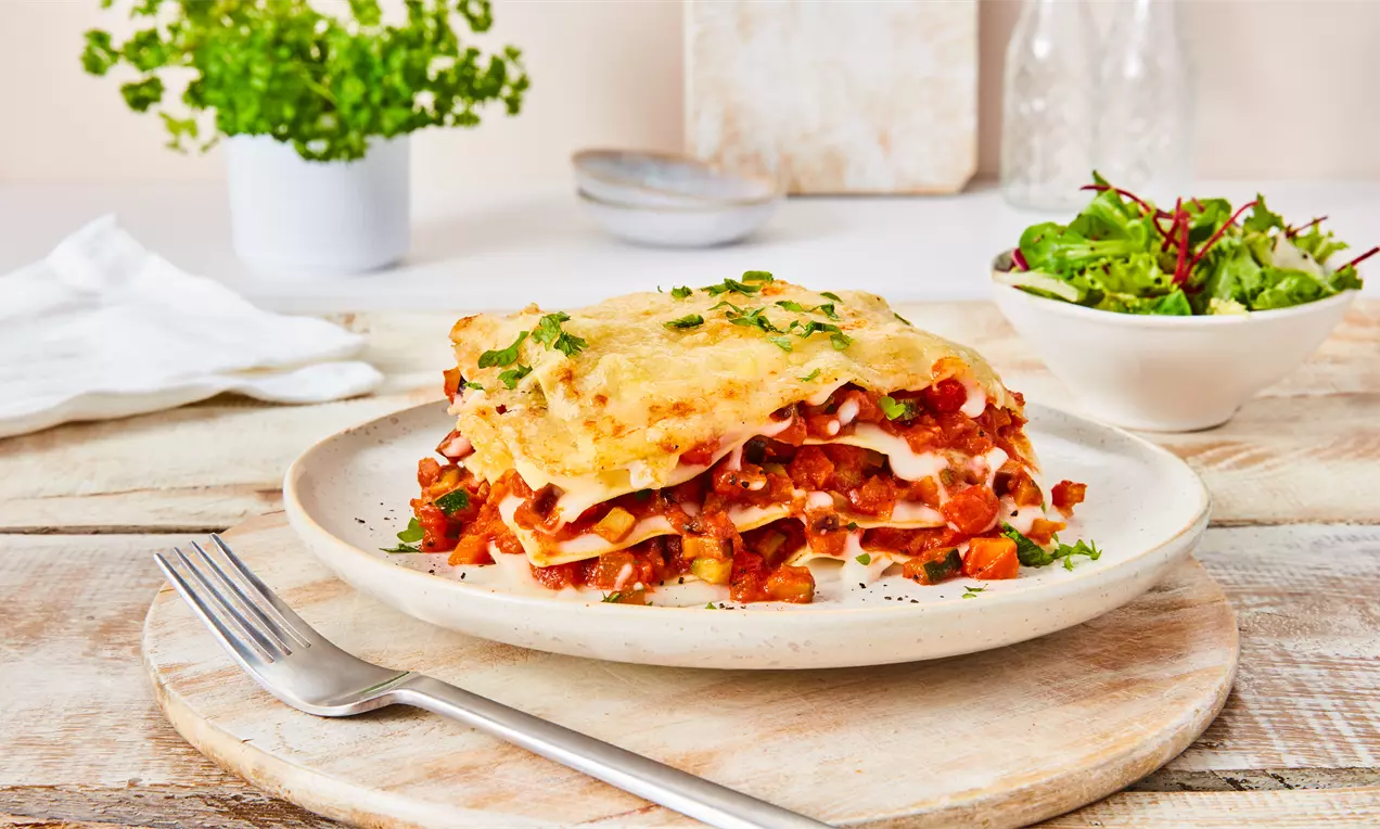 Vegane Gemüse-Lasagne mit geschichteter Tomatensauce, Bechamel, bunten Zucchini und Paprika, goldbraun und mit Basilikum