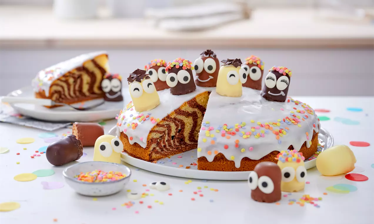 Zebrakuchen mit weißem Zuckerguss, bunten Zuckerstreuseln und Schokodeko in Kinderoptik, serviert auf einer weißen Platte