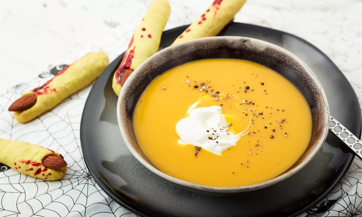 Kürbissuppe in einem dunklen, rundlichen Teller mit Sahnehäubchen und Pfeffer, arrangiert mit gruseligen Hexenfinger-Keksen auf Netz-Deckchen