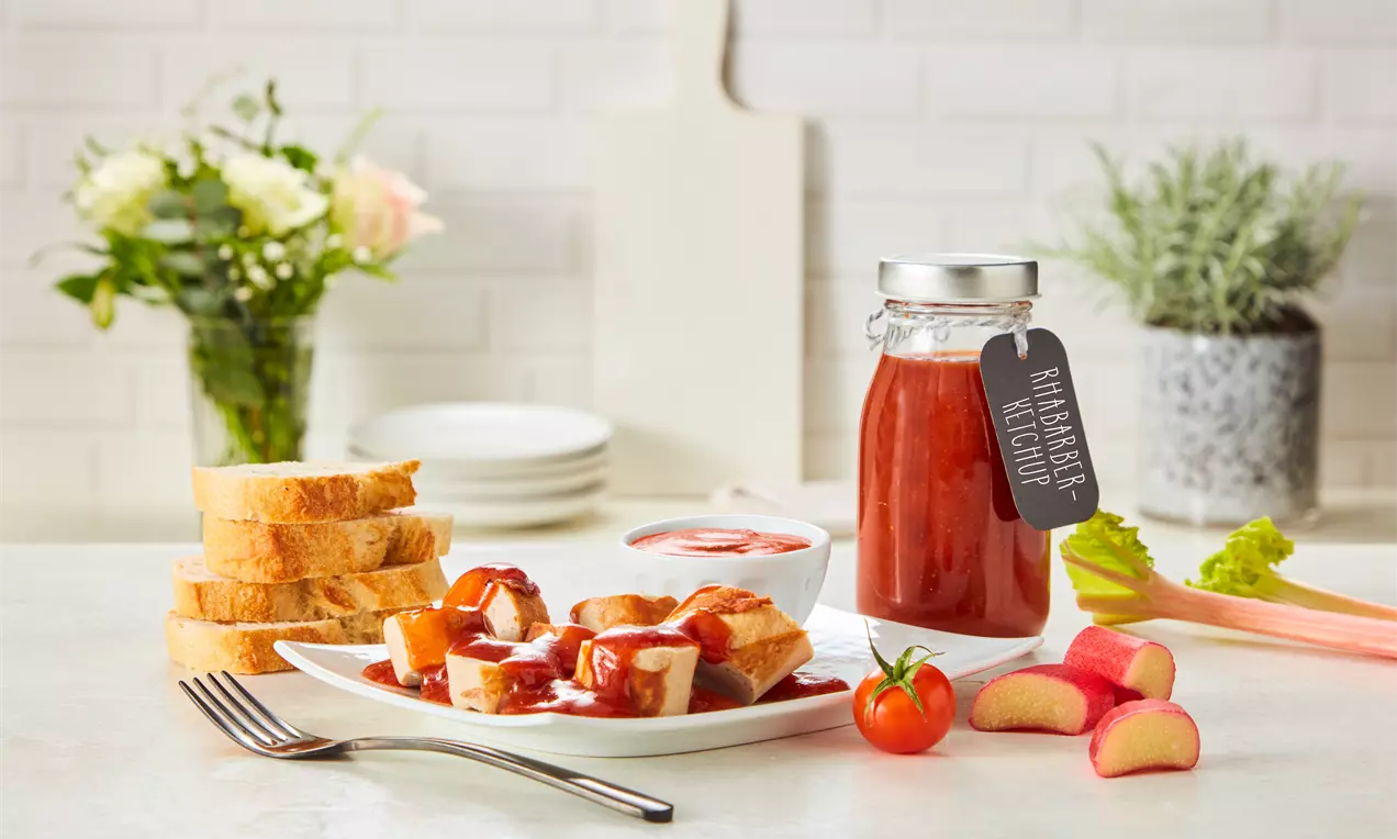 Rhabarberketchup in einer Glasflasche mit Etikett, umgeben von frischen Rhabarberstücken, Kirschtomate, Baguettescheiben und Dip-Schälchen