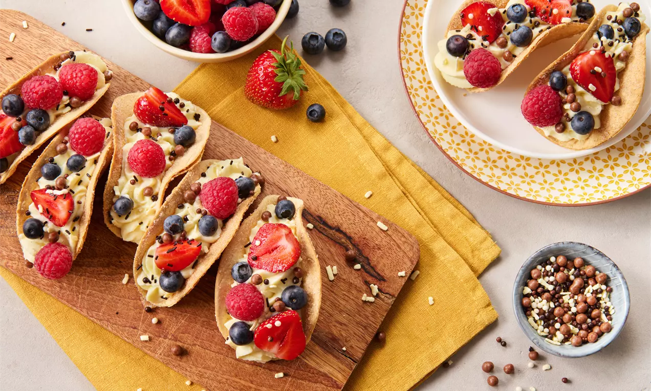 Knusprige Taccoschalen gefüllt mit heller Creme, dekoriert mit roten Erdbeeren, Himbeeren, blauen Heidelbeeren und Schokostreuseln