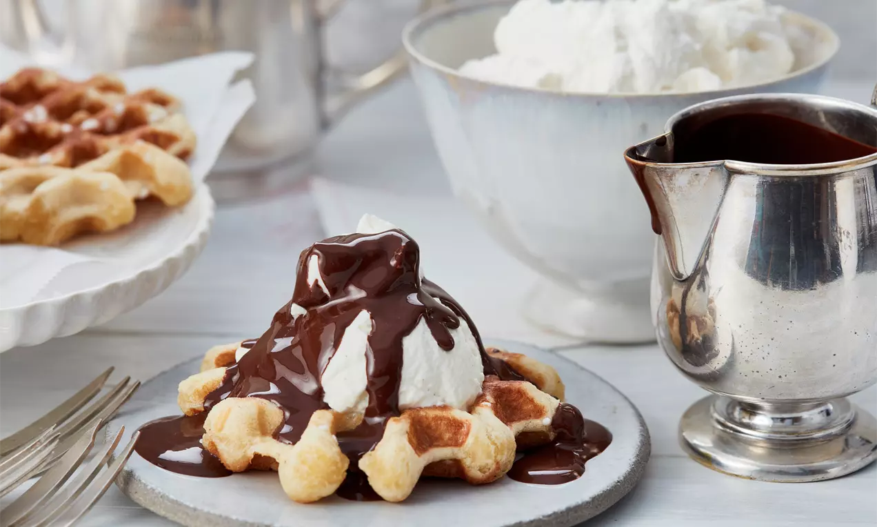 Goldbraune Lütticher Waffel auf grauem Teller, mit weißer Sahnehaube und glänzendem Schokoladenguss, umgeben von Sahneschüssel und Schokoladenkanne
