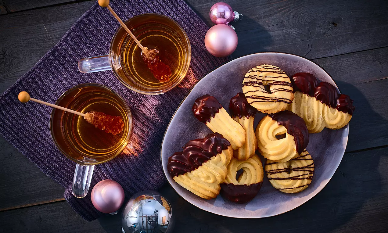 Veganes Spritzgebäck mit goldgelber Textur teils in dunkler Schokolade getaucht, angerichtet in einer Schale neben Teegläsern und Weihnachtskugeln