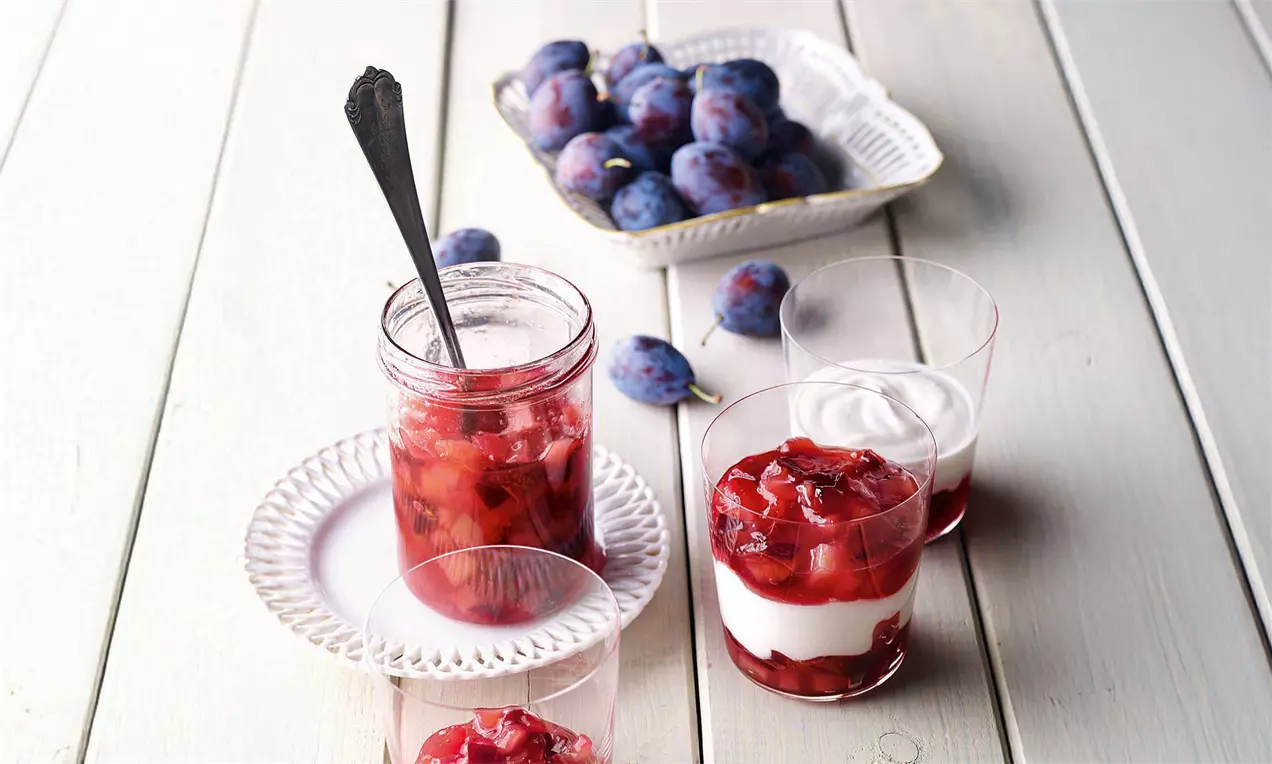 Rot-violetter Birnen-Zwetschgen-Kompott mit Zimt im Glas mit Löffel, daneben frische Zwetschgen und Dessertgläser mit Sahne auf hellem Untergrund