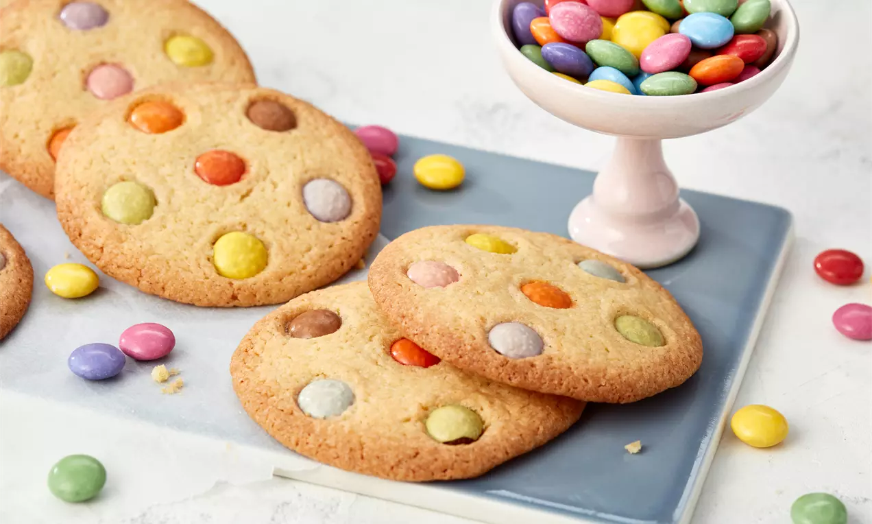Goldene, runde Cookies mit bunter Smarties-Dekoration auf einer blauen quadratischen Platte, daneben eine rosa Schale mit Smarties