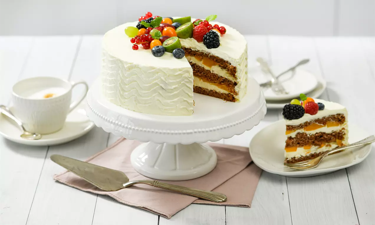 Mascarpone-Torte mit welliger weißer Creme, saftigem Schichtkuchen und buntem Obstbelag auf weißer Tortenplatte mit Kaffeegedeck