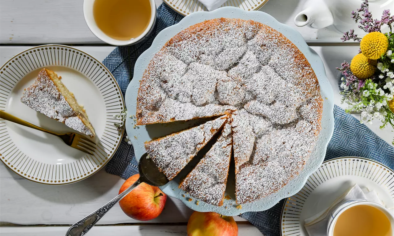 Glutenfreier Apfelkuchen mit goldbrauner Kruste und feinem Puderzucker, in Stücke geschnitten auf einer hellblauen Tortenplatte mit Äpfeln旁