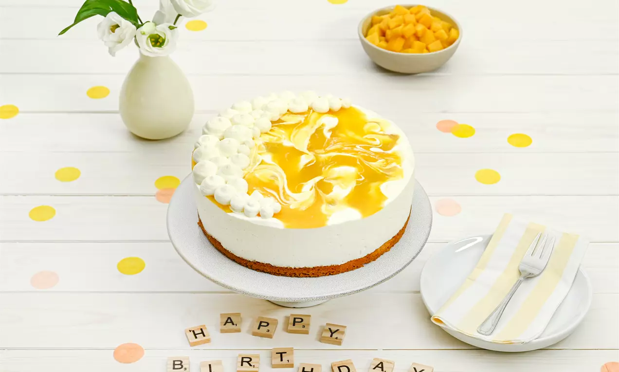 Käsesahne-Mango-Torte mit cremiger weißer Füllung auf braunem Boden, mit gelbem Mangospiegel und Sahnetupfen auf weißer Tortenplatte