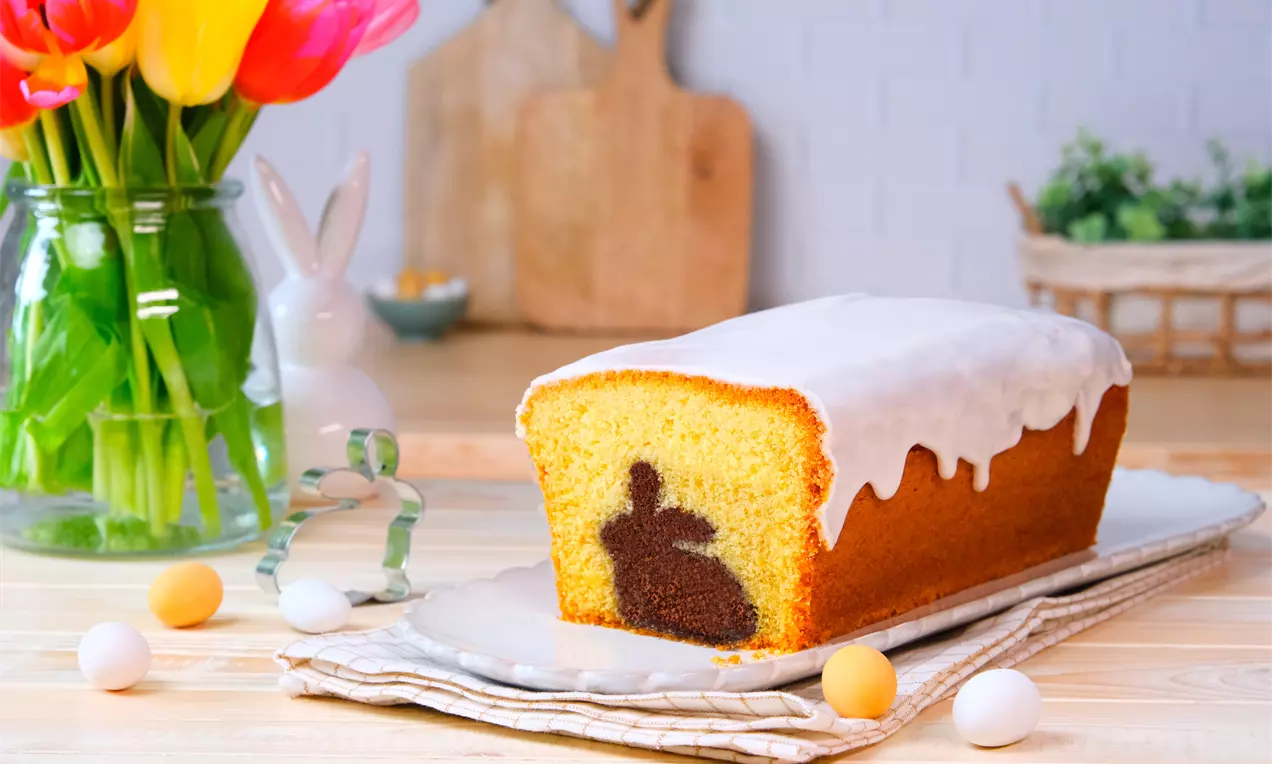 Kastenkuchen mit weißem Zuckerguss, im Anschnitt mit dunkler Hasensilhouette im Inneren; dekoriert mit bunten Mini‑Ostereiern, Tulpenstrauß und Küchenutensilien im Hintergrund.