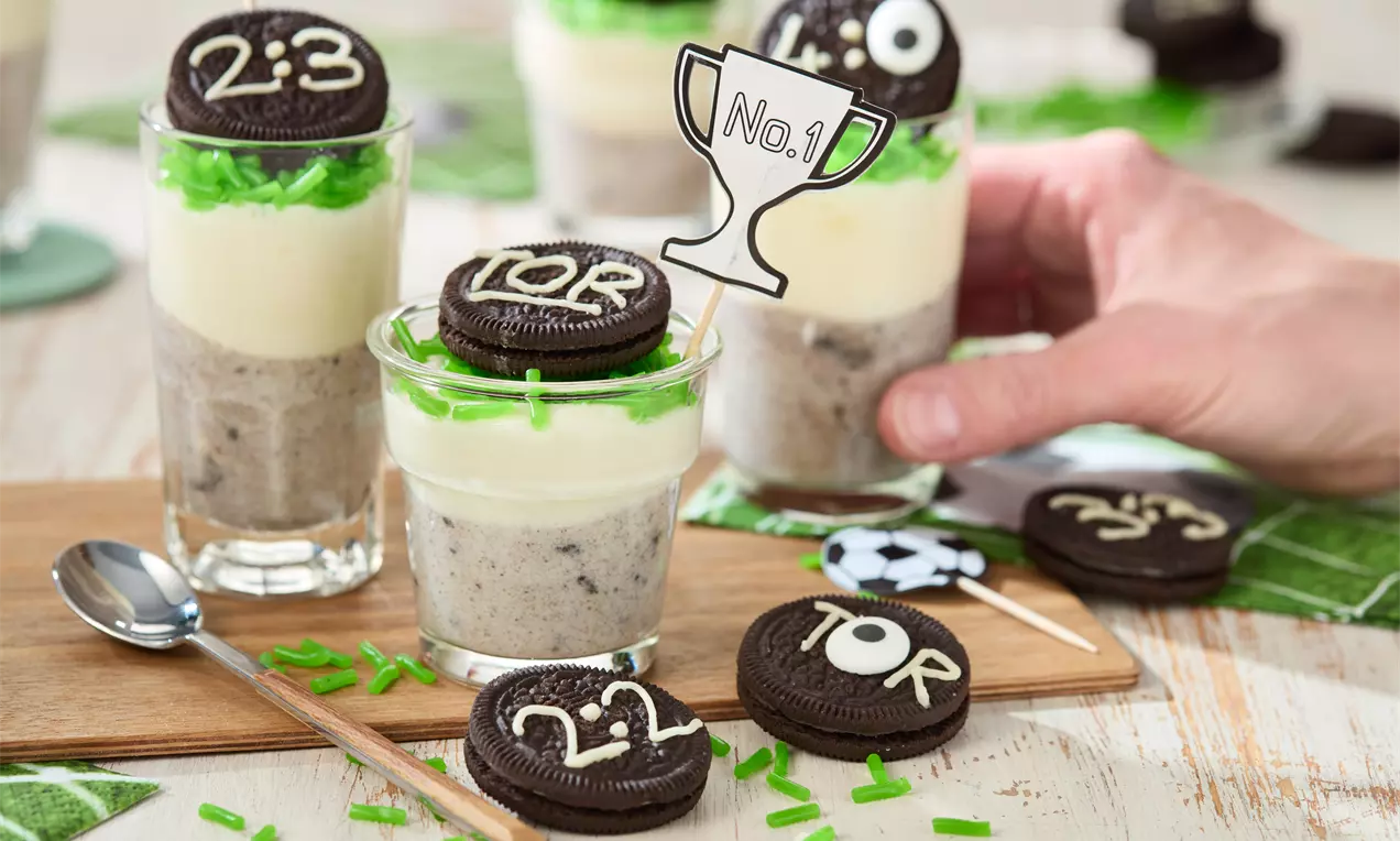Gläser mit Schichtdesserts aus weißer Creme und Oreobröseln, dekoriert mit grünem Zuckerdekor, Keksdeckeln und Fußball-Motiven auf Holzbrettern