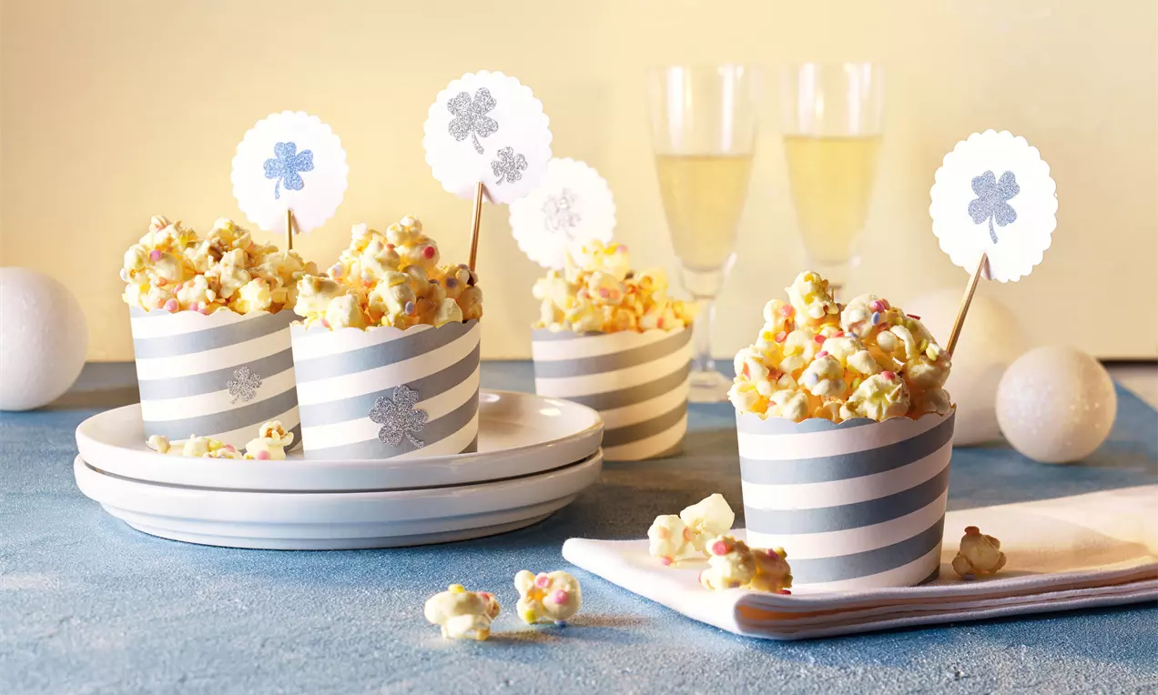 Weißes Schoko-Popcorn in gestreiften Papierbechern, mit bunten Streuseln verziert, arrangiert auf weißem Geschirr und blauer Tischdecke
