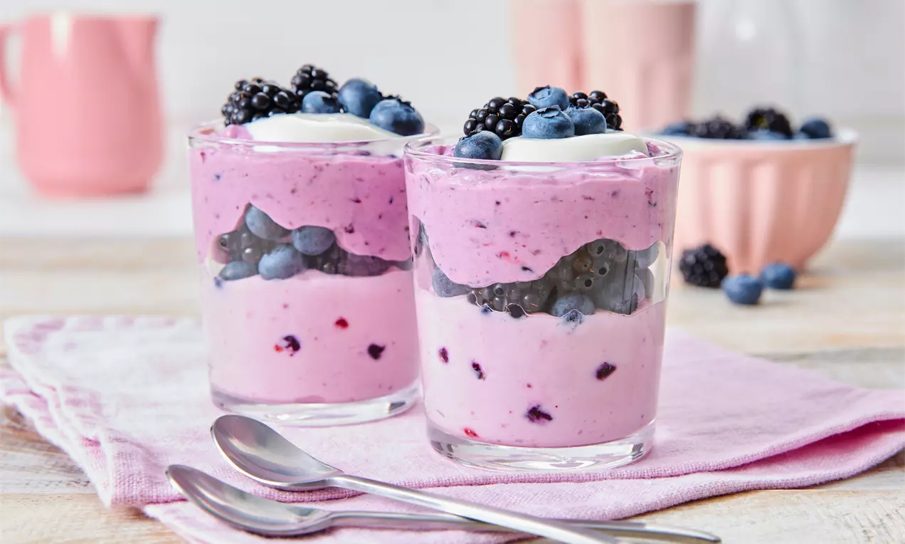Zwei durchsichtige Gläser mit rosafarbener Creme, Blaubeeren, Brombeeren und Sahnetupfen, auf einem rosa Tuch mit zwei Löffeln arrangiert