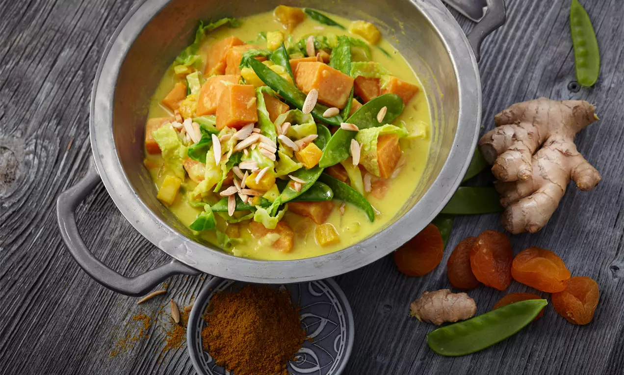 Wirsingcurry mit orangefarbenen Süßkartoffelwürfeln, grünen Zuckerschoten und hellgelber Soße in einer silbernen Servierschale