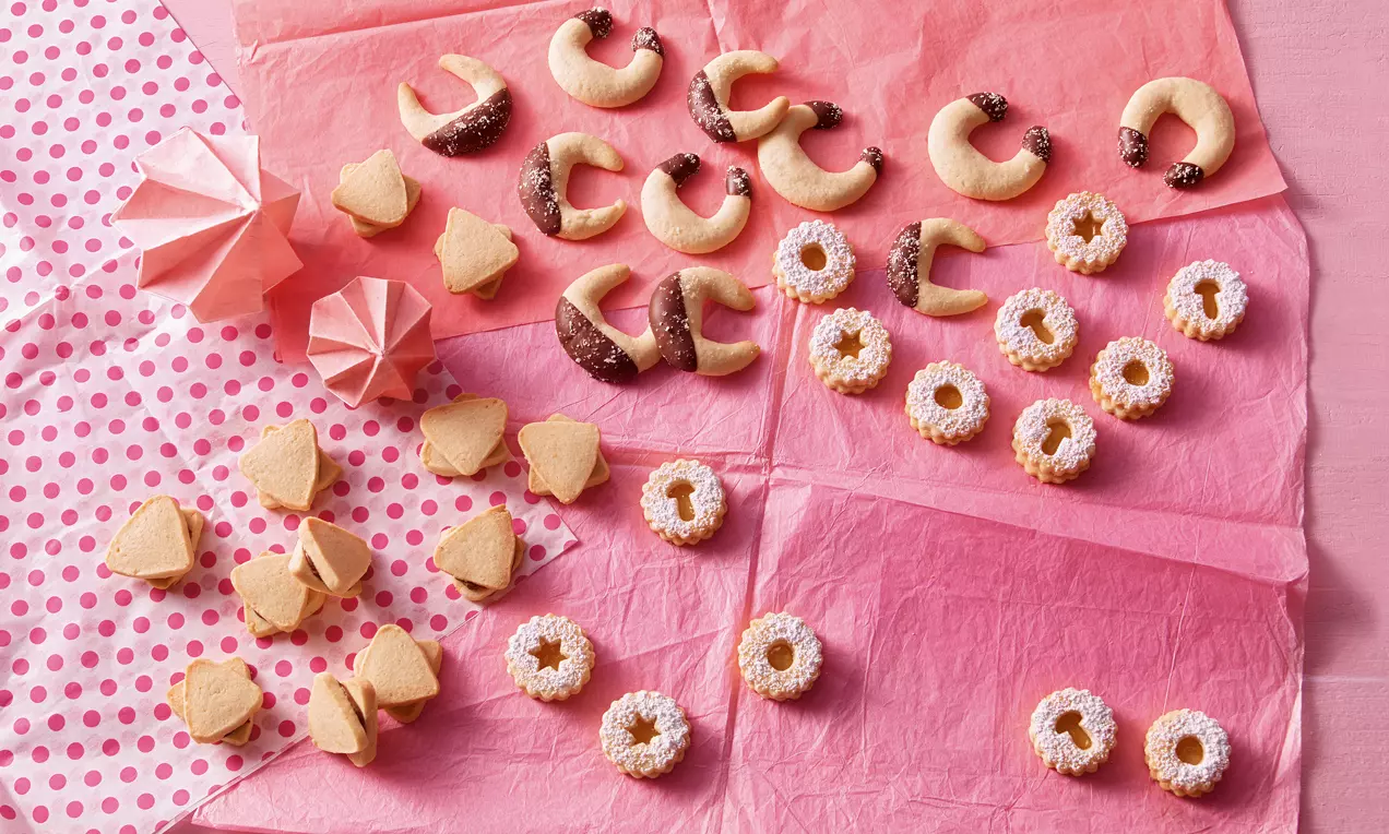 Vielfältiges Gebäck aus Vanillekipferl, Linzer-Plätzchen und Sternen, teils mit Puderzucker und Schokoladenglasur auf rosa Papier arrangiert