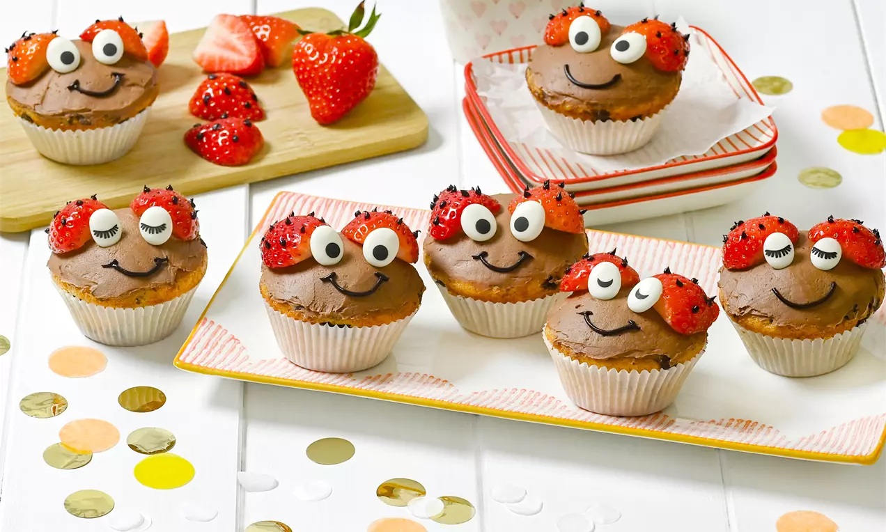 Marienkäfer-Muffins mit braunem Teig, glasierten Augen und Erdbeerhälften als Flügel auf bunten Tellern und Holzbrett angerichtet