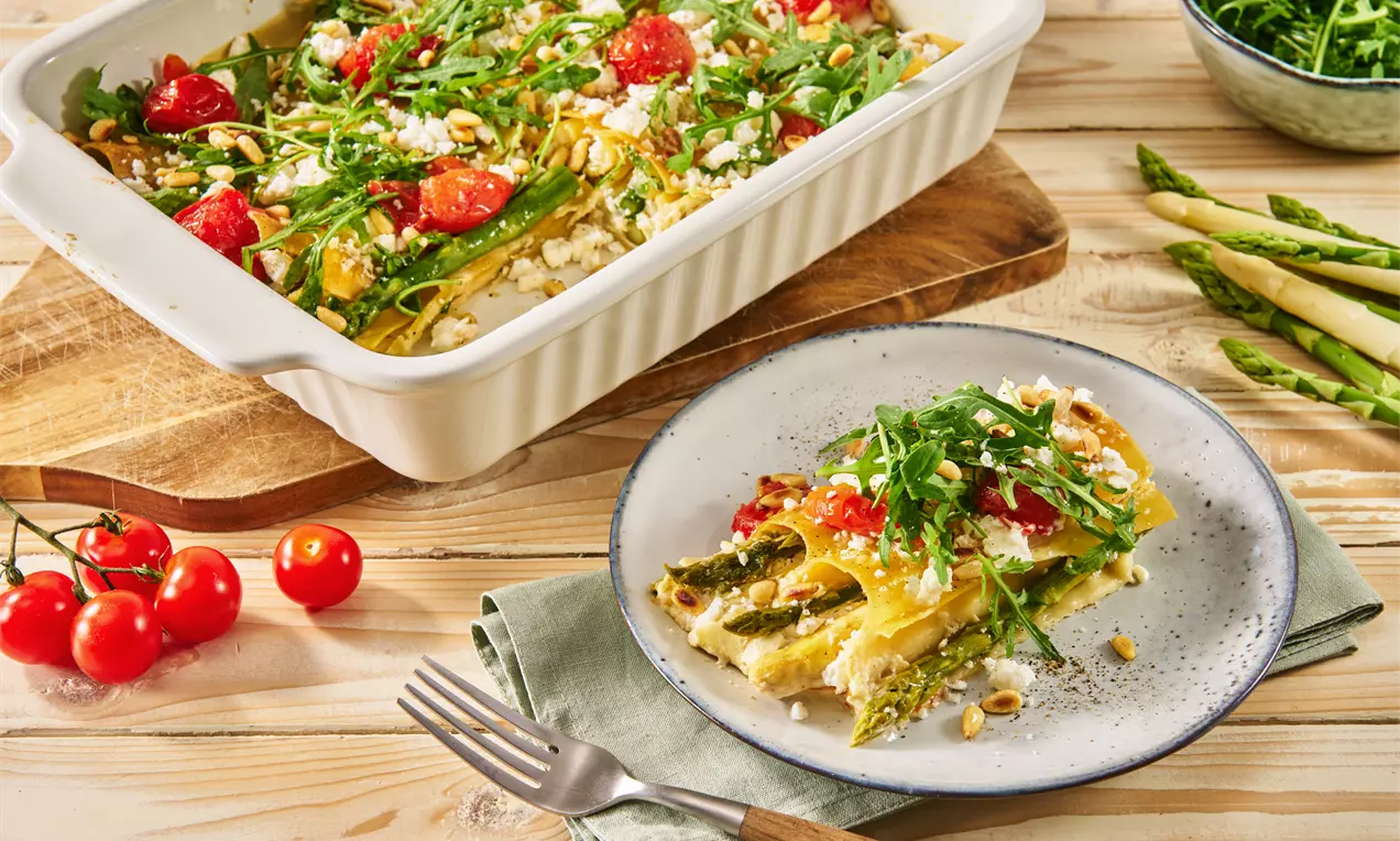 Goldgelb gebackene Cannelloni mit grünem Spargel, frischen Tomaten und Rucola, angerichtet in einer Keramikform und auf einem Teller mit Gabel