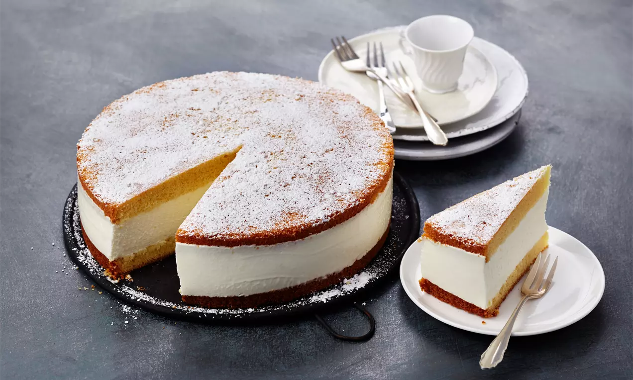 Käse-Sahne-Torte mit heller, cremiger Füllung, lockerem Baiser-Topping, frischen Himbeeren und Minzblatt auf weißem Tortenständer