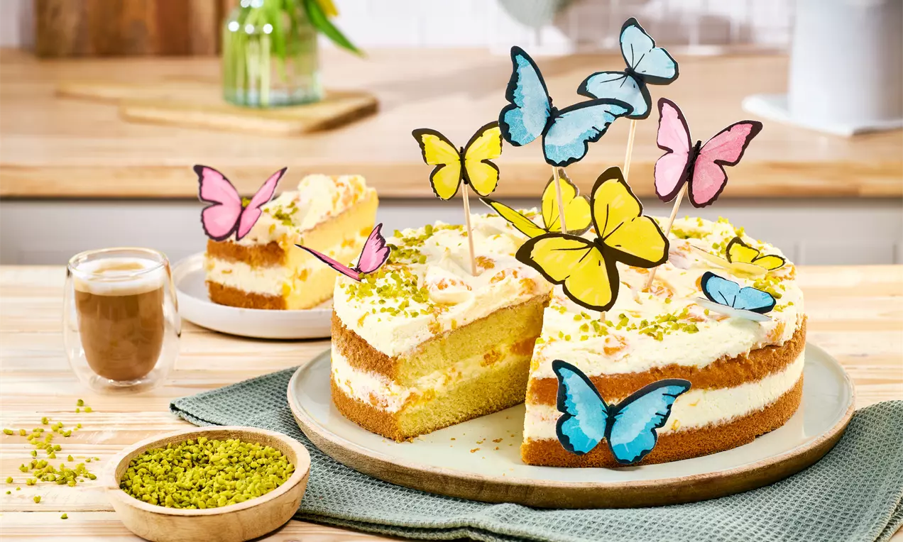 Frühlingstorte mit hellen Biskuitböden und Creme, bestreut mit Pistazien; dekoriert mit bunten Esspapier‑Schmetterlingen auf Holzstäbchen. Ein Stück ist herausgeschnitten und liegt leicht versetzt.
