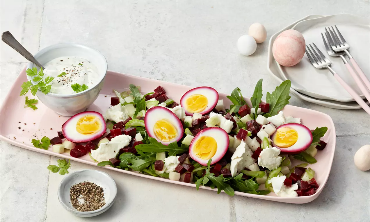 Halbierte rote Beete Eier mit gelbem Eigelb auf Rucola, Rote-Beete-Würfeln und Feta, angerichtet auf einer rosa Platte mit Dip-Schüssel