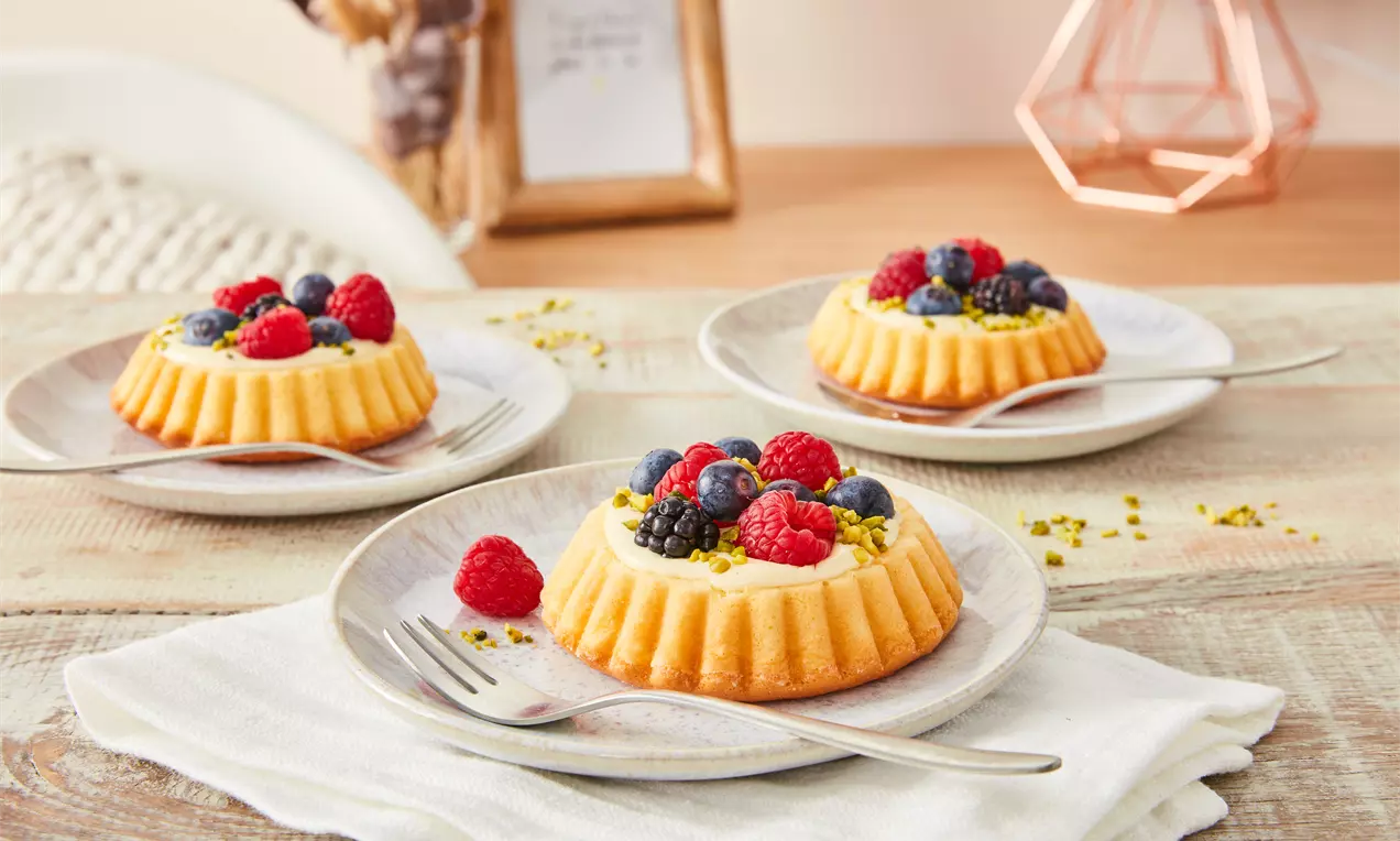 Kleine Obsttörtchen mit goldgelbem Mürbeteigboden, Sahnefüllung, frischen Beeren und Pistazien auf hellen Tellern mit Gabel angerichtet