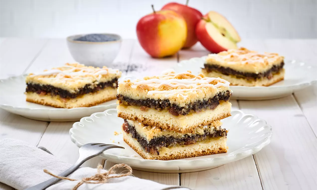 Sächsischer Mohnkuchen mit dunkler Mohnfüllung zwischen hellen Teigschichten und Streuseln auf weißen Tellern mit Äpfeln im Hintergrund