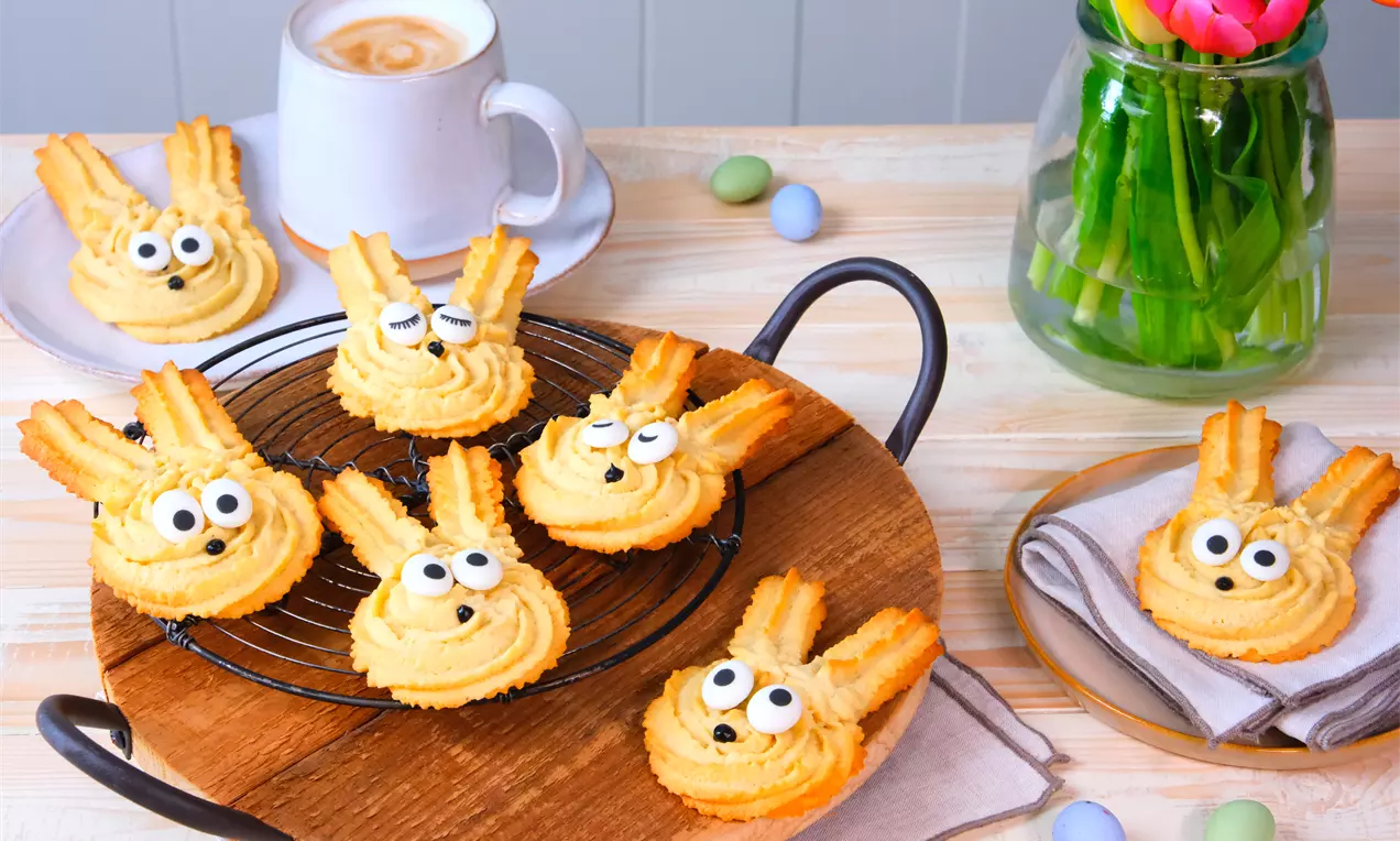 Helles, spritzgeformtes Ostergebäck in Hasenform mit Zuckeraugen auf schwarzem Gitter auf Holzbrett, umgeben von Dekoeiern und Tulpenstrauß