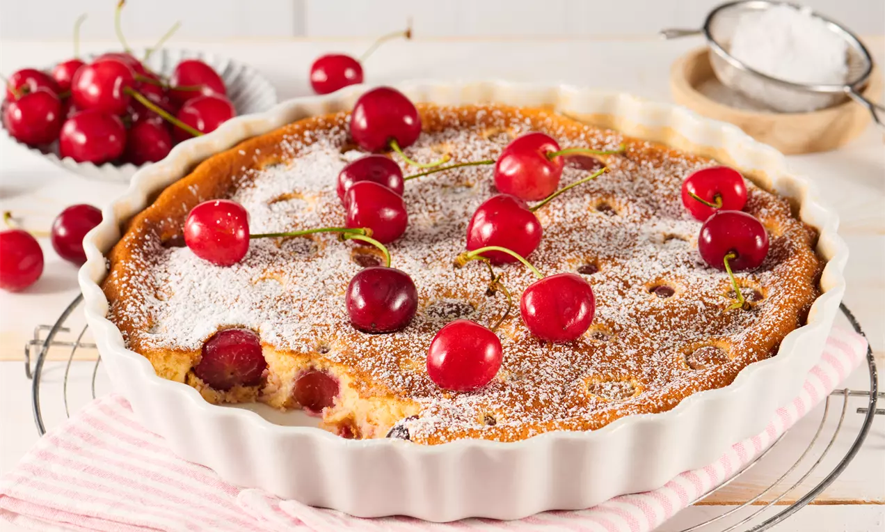 Goldbrauner Clafoutis mit frischen roten Kirschen und Puderzucker bestäubt in einer weißen Tarteform, auf einem gestreiften Tuch angerichtet