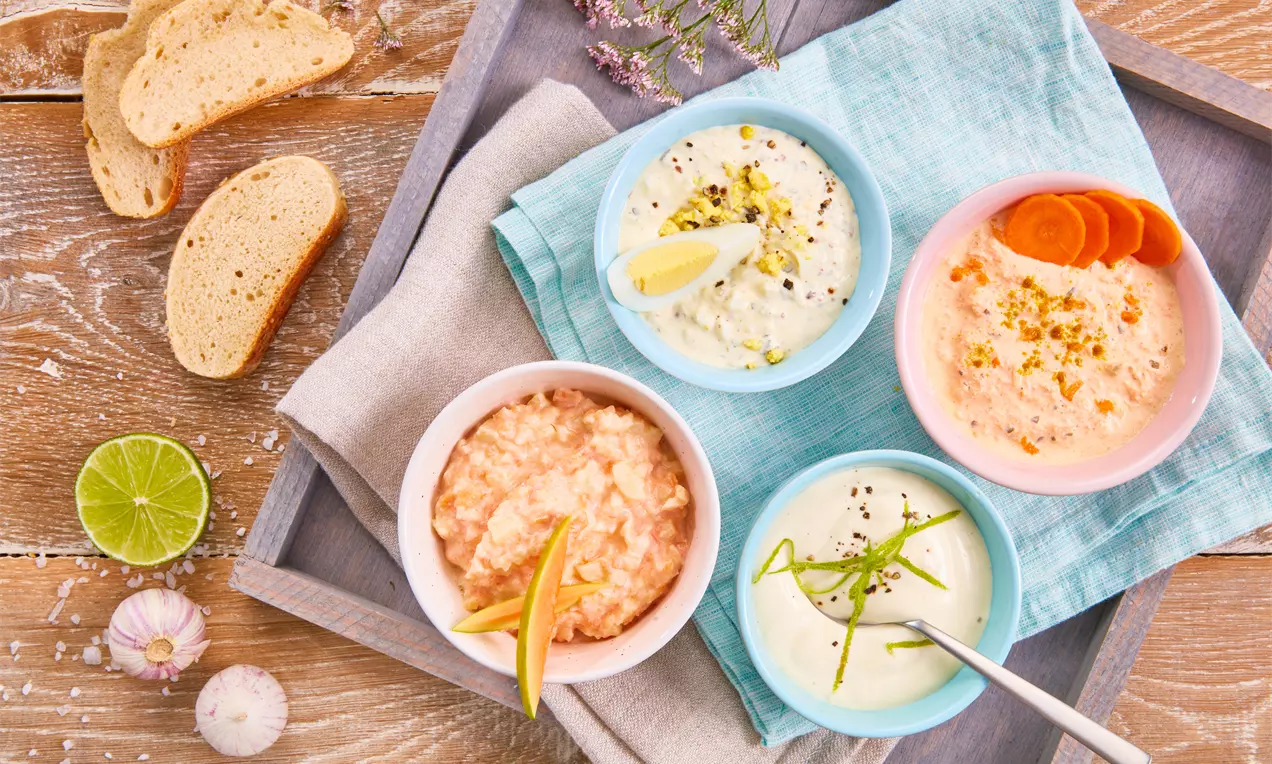 Vier bunte Schalen mit cremigen Crème-fraîche-Dips, dekoriert mit Gemüse und Kräutern auf blauem Stofftuch, flankiert von Brot und Limette