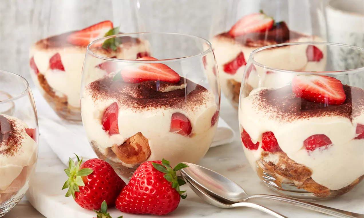 Erdbeer-Tiramisu in klaren Gläsern mit Schichten aus hellcremiger Mascarpone, rot-frischen Erdbeerstückchen und dunklem Kakao, dekoriert mit Erdbeeren und Löffel auf weißem Untergrund