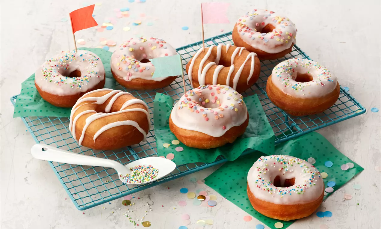 Vegane Donuts mit glänzender rosa Glasur, weißem Zuckerstreusel und Schokodrops auf rustikalem Holzbrett, einige gestapelt