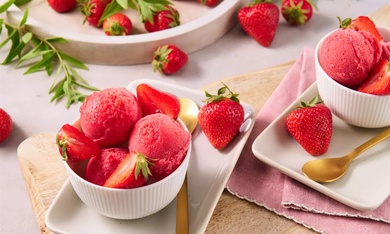 Knallrotes Erdbeersorbet in weißen Schalen, garniert mit frischen Erdbeeren und halbierten Früchten, auf hellen Tellern mit goldenen Löffeln arrangiert